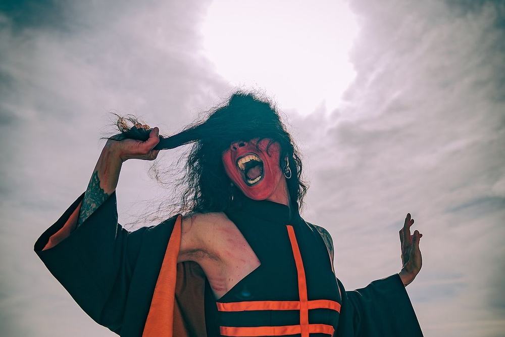 Dalila Kayros con maschera rossa e kimono nero e arancio, in controluce contro il sole sul mare
