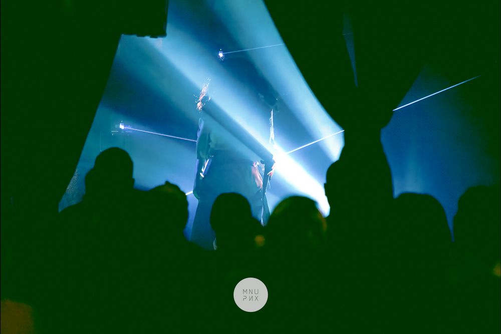 Palco avvolto da fasci di luce blu e laser, il pubblico in controluce
