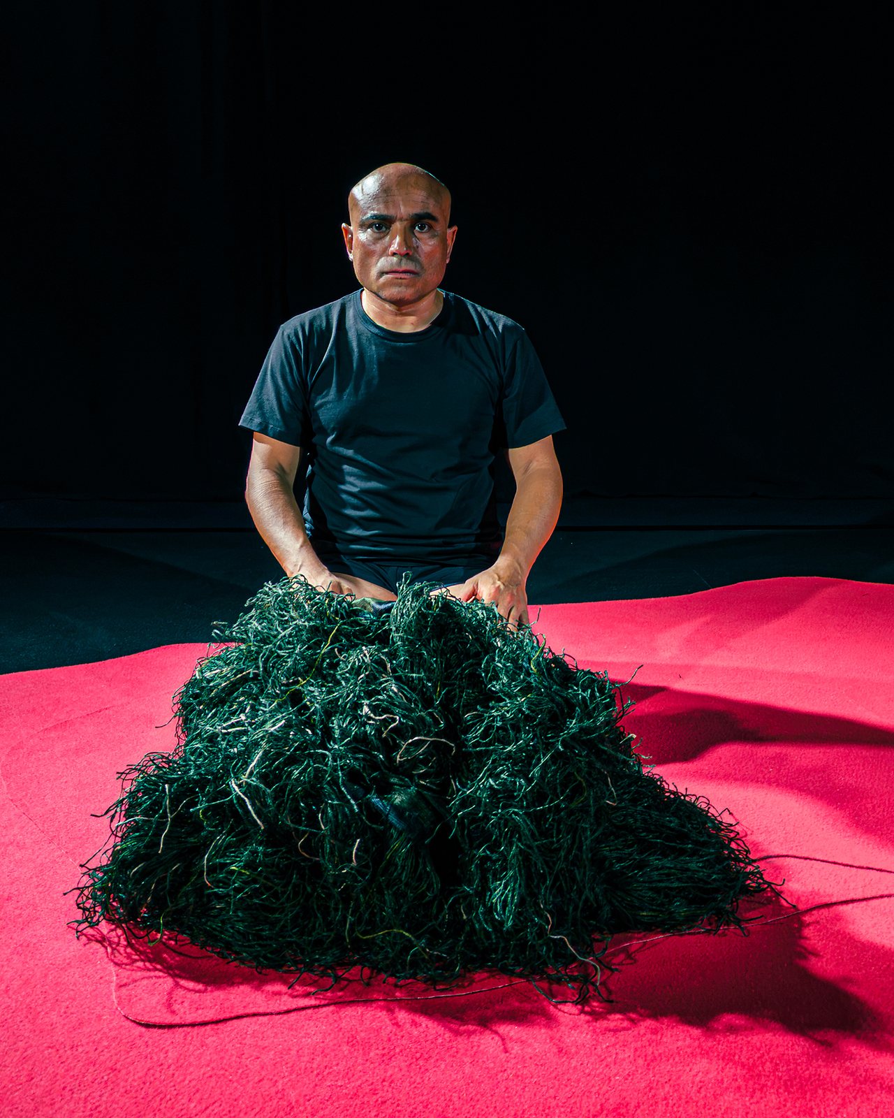 Performer kneeling behind a mass of dark green fibres on a red stage floor