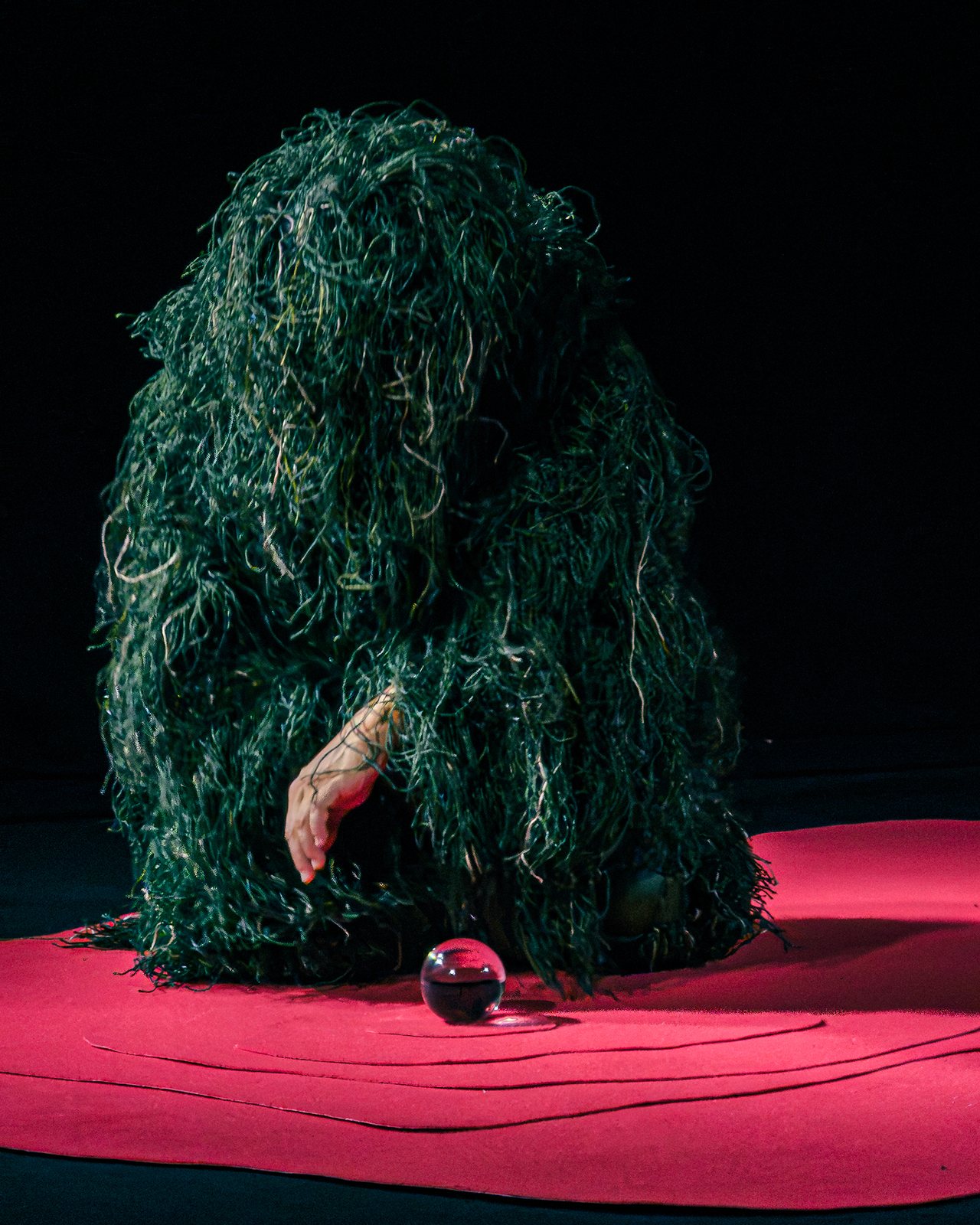 Figure entirely covered in dark green fibres crouching beside a red sphere